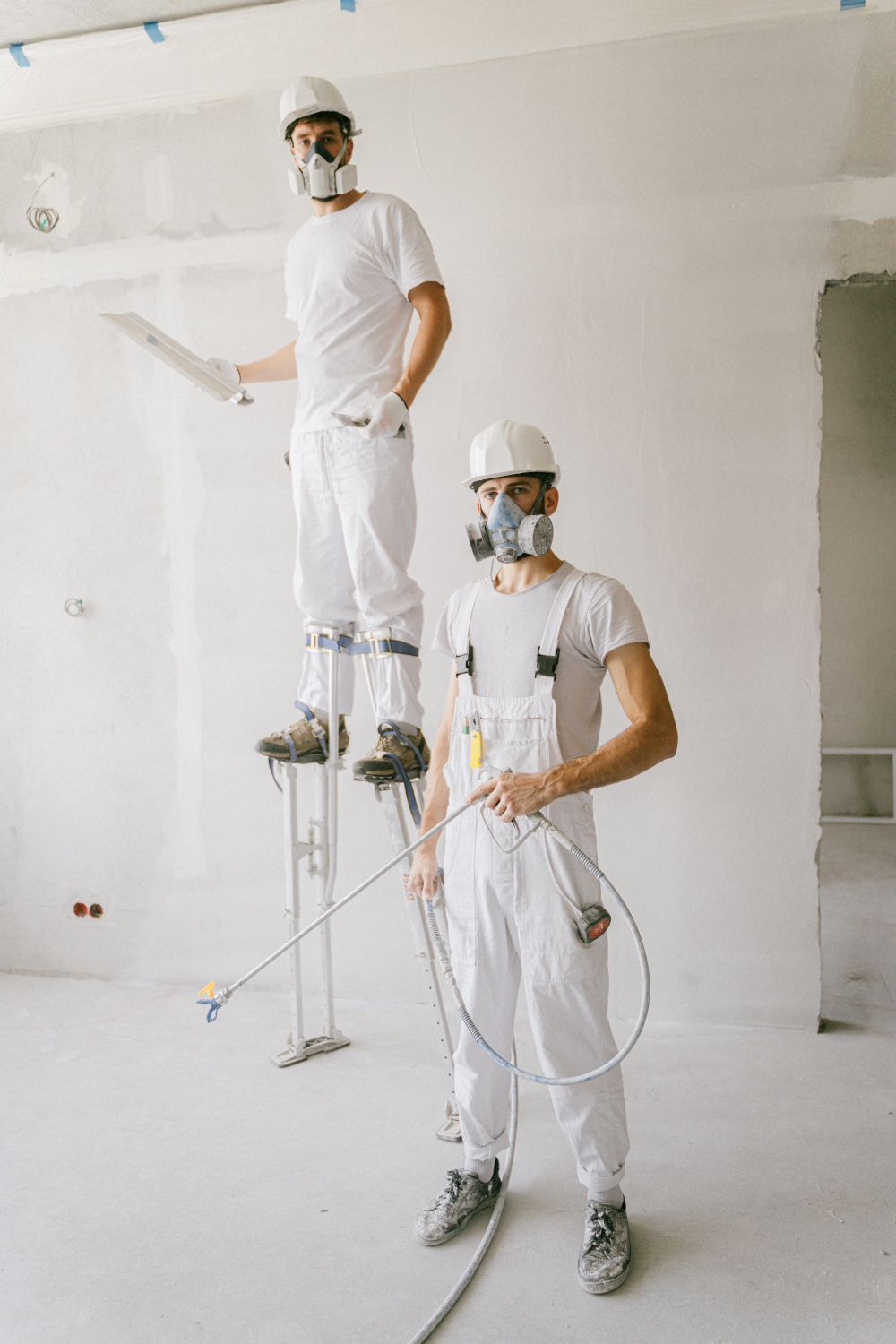 Two painters in white coveralls and respirators, one on stilts, prepping a room for spraying