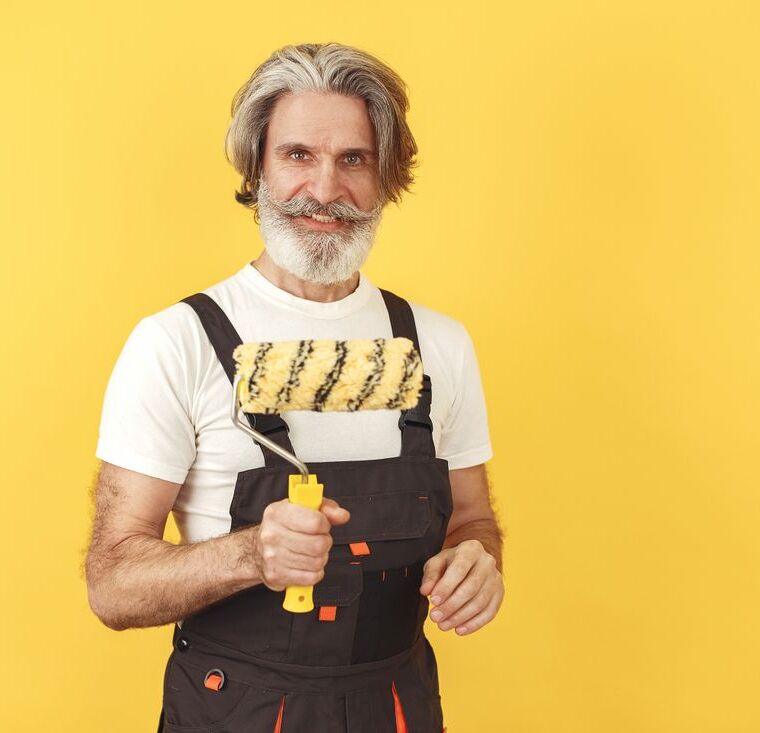 Dale Hartwell, gray-haired and bearded, holding a paint roller in work overalls