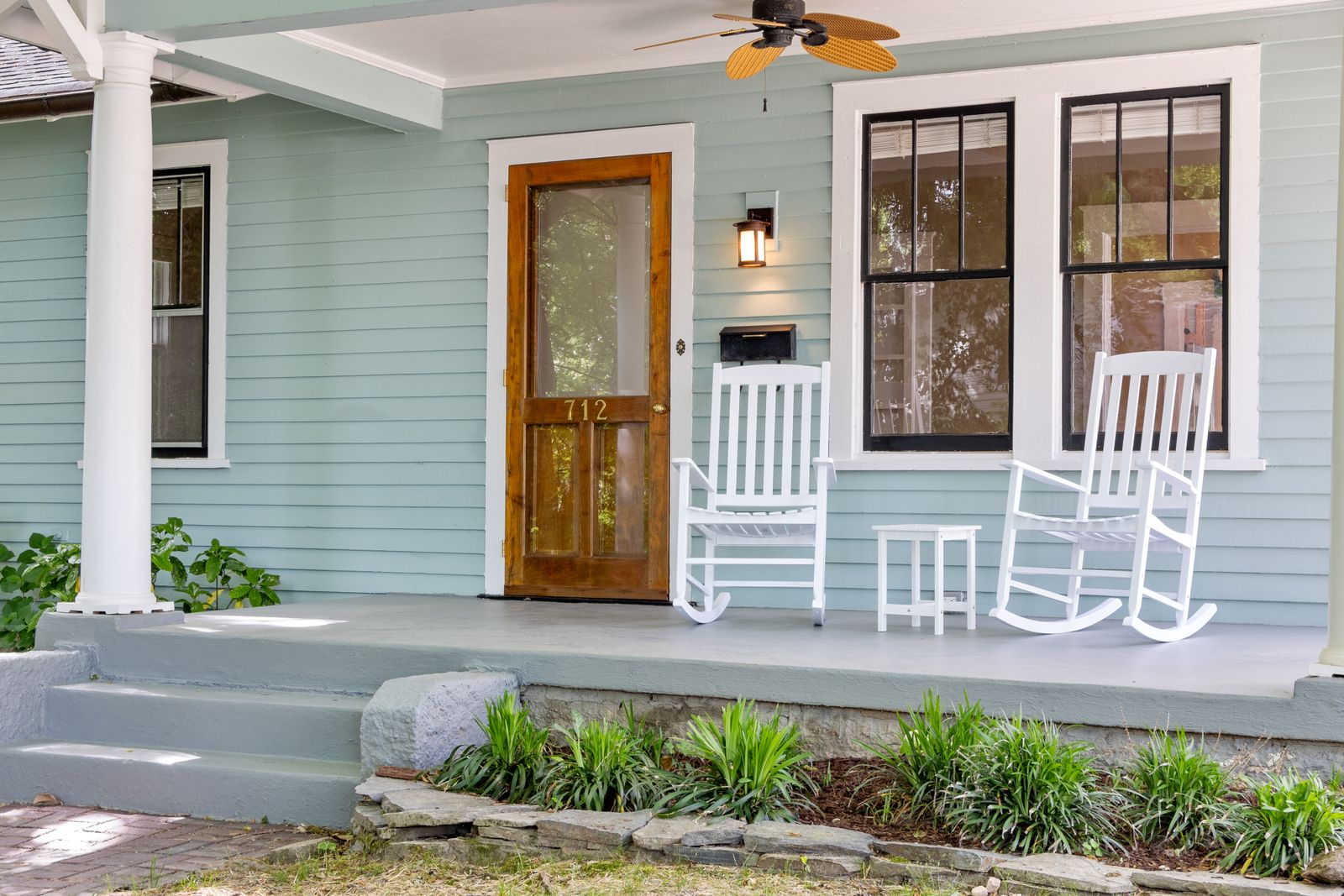 A pale blue house with white rocking chairs on the front porch and a freshly finished wood front door